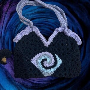 May include: Handmade crochet tote bag in navy blue with a light purple spiral design on the front. The bag has a light purple trim and handles. The background is a dark blue and purple gradient.