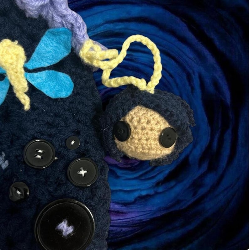 May include: A handmade crochet doll with button eyes and dark blue hair, attached to a dark blue crochet item with large black buttons. The doll has a tan face and is connected by a yellow crochet loop. Blue and yellow dragonfly accents are also visible.