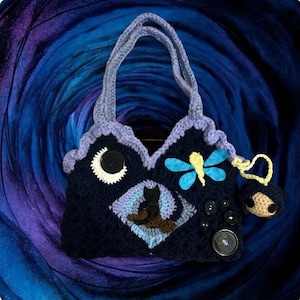May include: Handmade crochet handbag in navy blue with a whimsical design. Features a crescent moon, a black cat, and a dragonfly. Adorned with decorative buttons and a small charm. The bag has a lavender trim and handles.
