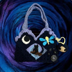 May include: Handmade crochet handbag in navy blue with a whimsical design. Features a crescent moon, a black cat, and a dragonfly. Adorned with decorative buttons and a small charm. The bag has a lavender trim and handles.