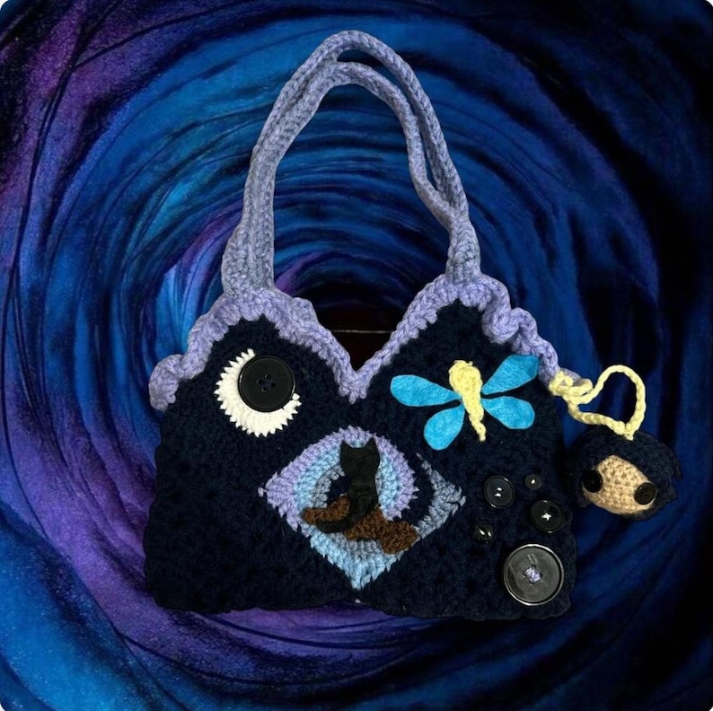 May include: Handmade crochet handbag in navy blue with a whimsical design. Features a crescent moon, a black cat, and a dragonfly. Adorned with decorative buttons and a small charm. The bag has a lavender trim and handles.