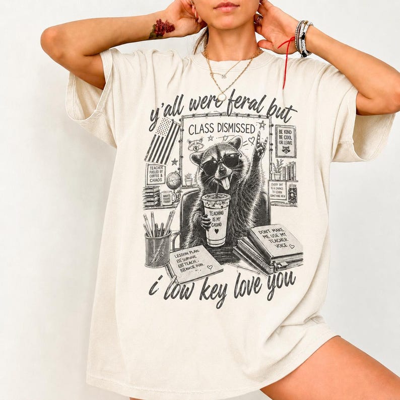 Op de afbeelding: Cr&egrave;mekleurig T-shirt met een afbeelding van een wasbeer met een zonnebril, die een beker vasthoudt en het vredesteken maakt. Het shirt heeft de tekst "y'all were feral but class dismissed" en "i low key love you."
