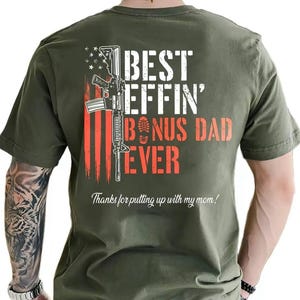 May include: Olive green t-shirt with a graphic design featuring a rifle, the American flag, and the words "BEST EFFIN' BONUS DAD EVER." The text "Thanks for putting up with my mom!" is also included.
