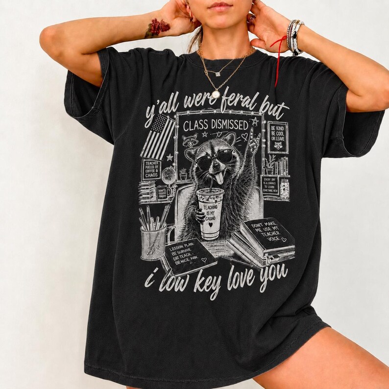 Op de afbeelding: Zwart grafisch t-shirt met een wasbeer met zonnebril, die een koffiebeker vasthoudt en het vredesteken maakt. De tekst "y'all were feral but CLASS DISMISSED" en "i low key love you" is inbegrepen.