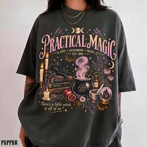 May include: A charcoal gray t-shirt featuring a graphic print with the text "PRACTICAL MAGIC," "LOVE + SISTERHOOD + MAGIC," "EST. 1995," and "There's a little witch in all of us." The design includes a black cat, books, candles, a cauldron, and flowers.