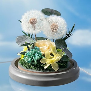 May include: A decorative floral arrangement featuring two large white dandelions, a cream-colored rose, and various green and yellow accent flowers. The arrangement is set in a gray, dome-shaped container with a moss base, against a blue sky backdrop.