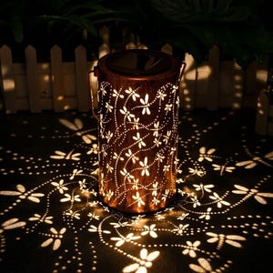Puede incluir: Una linterna solar de color bronce con un diseño de libélulas. La linterna proyecta sombras con forma de libélula. Tiene un asa y un panel solar. Ideal para exteriores, crea un efecto de luz decorativo en jardines o patios.