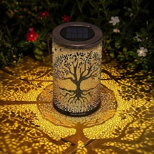 Puede incluir: Una linterna solar cilíndrica con diseño de árbol de la vida. La linterna es de metal con acabado bronce. El diseño del árbol está recortado, permitiendo que la luz brille a través y proyecte una sombra en forma de árbol. La parte superior tiene un panel solar. Se lee "SOLAR LANTERN".