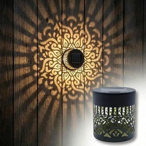 May include: Solar-powered outdoor light with a decorative, patterned design. The light casts an intricate pattern on a wooden surface. The black cylindrical lamp has a solar panel on top and a cut-out design that allows light to shine through.