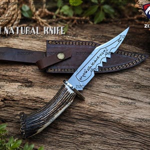 May include: A fixed-blade knife with a curved, textured brown antler handle and a silver-colored steel blade featuring a serrated edge and etched runic symbols. The knife is accompanied by a brown leather sheath with visible stitching.