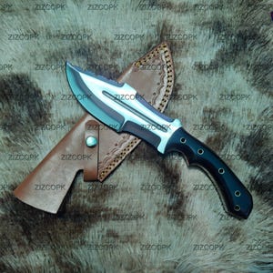 Puede incluir: Un cuchillo de caza con una hoja plateada y un mango negro descansa sobre una funda de cuero marrón. El cuchillo tiene una hoja curva y un mango resistente con una serie de pequeños agujeros. La funda también es marrón y tiene un patrón decorativo.