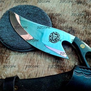 May include: A fixed-blade knife with a polished, curved steel blade featuring a circular emblem of a skull. The handle is black with a cutout design and a small circular inlay. A black leather sheath is positioned below the knife blade.