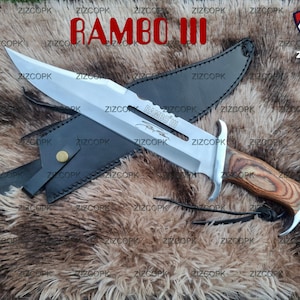 Può includere: Un coltello grande, color argento, con manico in legno e fodero in pelle nera. La lama è incisa con "RAMBO III" e una firma. Il coltello è esposto su una superficie marrone strutturata.
