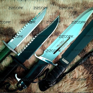 Peut inclure: Quatre couteaux de survie avec des lames dentelées et divers modèles de manches. L'un des couteaux a un manche vert, tandis qu'un autre porte le mot "RAMBO" sur la lame. Les couteaux sont exposés sur une surface brune texturée.