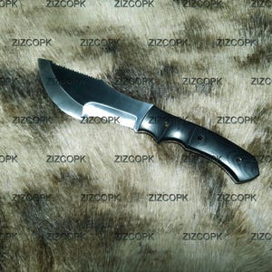 May include: A black and silver hunting knife with a serrated top edge and a curved blade. The handle is black and appears to be made of wood. The knife is displayed on a textured, brown and gray surface.