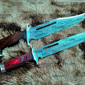 May include: Two Damascus steel knives with wooden and red handles. The blades have a unique pattern and a serrated edge near the handle. The knives are displayed on a textured, brown surface.
