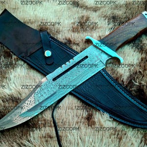 May include: A large, ornate knife with a patterned blade and wooden handle, resting on a black leather sheath. The knife has a decorative design and a turquoise accent. The sheath is embossed with a crocodile pattern.