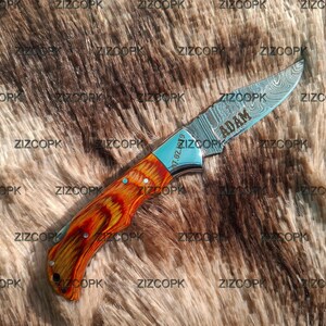 May include: A Damascus steel folding knife with a wooden handle. The blade is etched with the name "ADAM". The handle is a mix of orange and brown tones. The knife has a silver bolster and a date is engraved on the blade.
