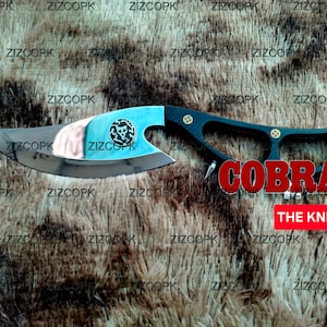 Puede incluir: Un cuchillo personalizado con una hoja curva y un mango negro. La hoja tiene una sección azul claro con un logotipo de calavera. La palabra "COBRA" se muestra en rojo, y el texto "THE KNIFE" está debajo.