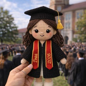 May include: A crocheted doll wearing a black graduation gown and cap with a gold tassel. The doll has brown hair and a red and gold sash that reads "Linaa 2028". The doll is held in front of a crowd of people in graduation attire.