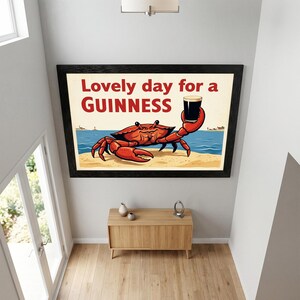May include: Framed art print featuring a cartoon crab holding a pint of Guinness. The crab is red, set against a beach scene with blue water and a light tan sand. The text "Lovely day for a Guinness" is displayed in red.