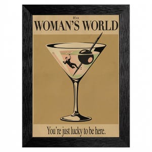 May include: Framed art print with a martini glass design. The print features the text "it's a WOMAN'S WORLD" above the glass and "You're just lucky to be here" below. The martini glass has an olive and a figure falling into the drink.
