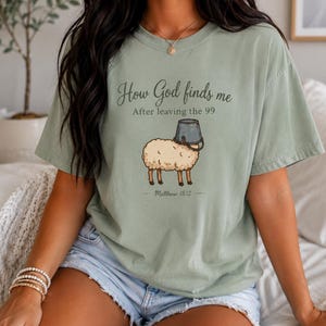 May include: A sage green short-sleeve t-shirt featuring a graphic of a sheep with a metal bucket on its head. The text reads: How God finds me After leaving the 99, and below the sheep, Matthew 18:12. The shirt has a crew neckline.