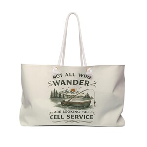 May include: Cream-colored tote bag with white rope handles. The bag features a graphic of a boat on a lake with mountains and the text "NOT ALL WHO WANDER ARE LOOKING FOR CELL SERVICE."