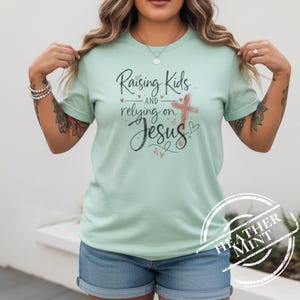 Może przedstawiać: Jasnozielona koszulka z napisem "Raising Kids and relying on Jesus" w czarnym skrypcie. Różowy krzyż i serca również znajdują się na koszulce. Koszulka jest połączona z niebieskimi szortami dżinsowymi.