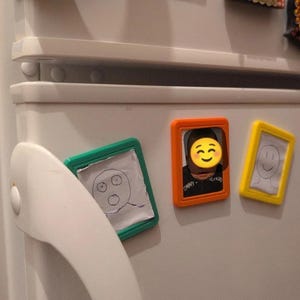 May include: Three rectangular magnetic picture frames in teal, orange, and yellow. The teal frame holds a hand-drawn sketch of a surprised face. The orange frame contains a photograph of a person with a yellow smiley face emoji overlay. The yellow frame displays a hand-drawn sketch of a simple smiling face.