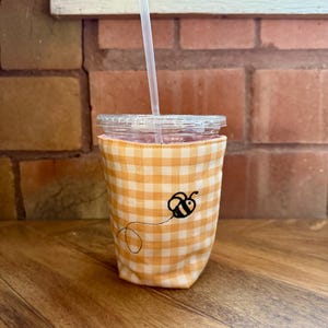Op de afbeelding: Een doorzichtige plastic beker met een rietje, in een geel en wit geruite stoffen hoes. De hoes heeft een zwart geborduurd bijenontwerp. De beker staat op een houten oppervlak.