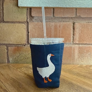 Op de afbeelding: Een marineblauwe drankhoes met een wit ganzenontwerp en oranje accenten. De hoes is gewikkeld rond een helder glas met een rietje. De achtergrond omvat een bakstenen muur en een houten oppervlak.