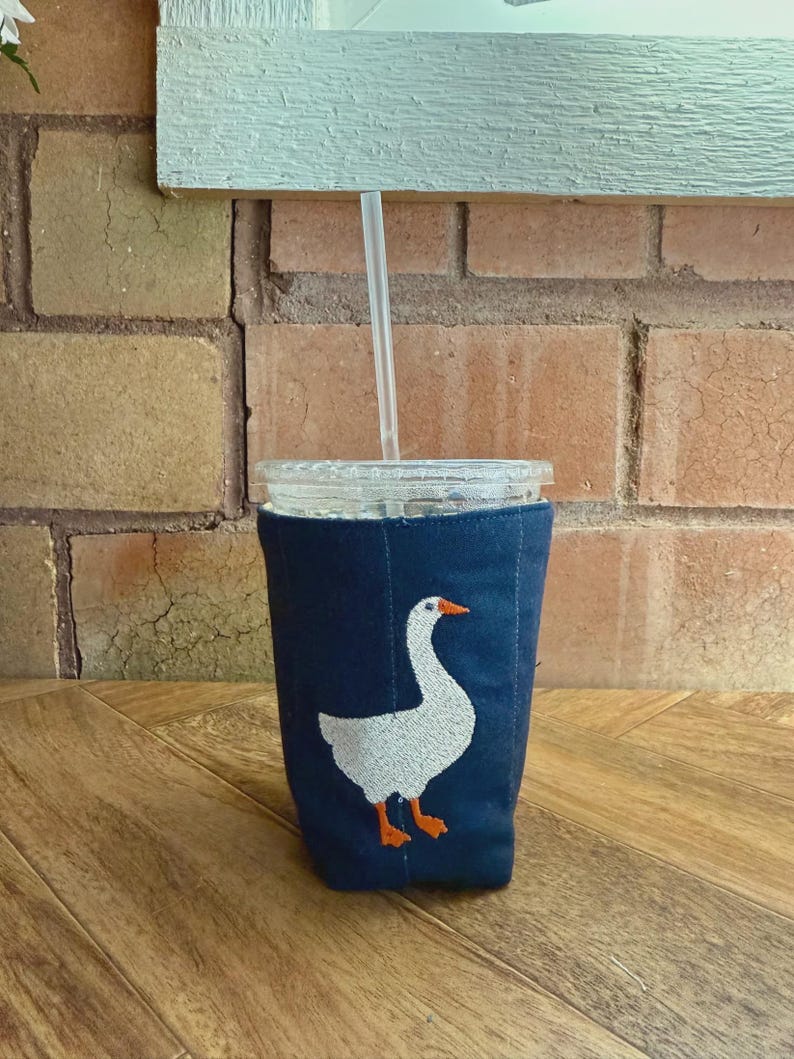 Op de afbeelding: Een marineblauwe drankhoes met een wit ganzenontwerp en oranje accenten. De hoes is gewikkeld rond een helder glas met een rietje. De achtergrond omvat een bakstenen muur en een houten oppervlak.