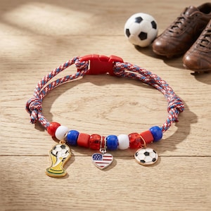 Op de afbeelding: Een armband van rood, wit en blauw gevlochten koord met een rode plastic gesp. Het bevat kralen in rood, wit en blauw, en drie metalen bedels: een goudkleurige trofee, een hartje met de Amerikaanse vlag en een zwart-witte voetbal.