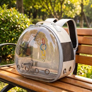 May include: A white pet carrier backpack with a large, clear, semi-spherical front window. The interior features a wooden perch, a hanging toy with red, blue, and yellow rings, and a small metal bell. It includes side mesh panels and metal grommets.