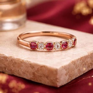 May include: A rose gold ring featuring alternating red gemstones and clear, round-cut stones. The ring is set on a light-colored stone surface, with a blurred background of red fabric and a golden object.