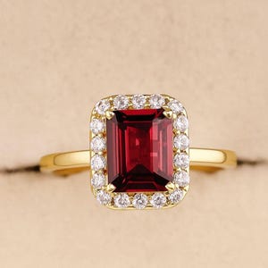 May include: A gold ring with a rectangular, deep red gemstone, encircled by a halo of small, clear stones. The ring's band is plain gold. The gemstone is the main feature, with a rich, saturated colour.
