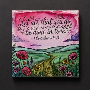 Puede incluir: Un azulejo decorativo cuadrado con un paisaje estilo acuarela que muestra un camino de tierra, colinas verdes y flores silvestres coloridas. El cielo es rosa y morado. El texto dice: Let all that you do be done in love. -1 Corinthians 16:14.