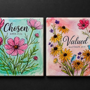 May include: Two square ceramic tiles featuring watercolor-style floral illustrations. The left tile has pink cosmos flowers on a teal background with the text Chosen JOHN 15:16. The right tile has yellow and pink flowers with blue birds and the text Valued MATTHEW 10:31.