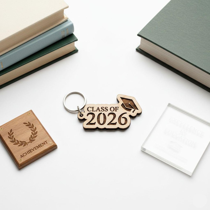 May include: A wooden keychain with the text "CLASS OF 2026" and a graduation cap design. Also, a wooden plaque with a laurel wreath and the word "ACHIEVEMENT" is visible. The items are on a white surface.