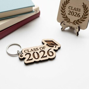 May include: Wooden keychain with the text "CLASS OF 2026" and a graduation cap design. The keychain is attached to a silver metal ring. A wooden coaster with "CLASS 2026" is also visible. The items are made of light-colored wood.