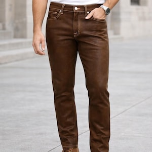 May include: Brown leather pants with visible stitching and a button closure. The pants are paired with brown leather boots and a white t-shirt. The pants have a classic five-pocket design.