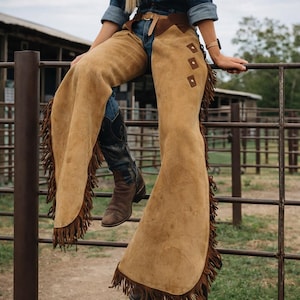 May include: Tan suede leather chaps with decorative square accents and fringe trim. The chaps are worn over blue jeans and brown cowboy boots. The chaps are a western wear item.