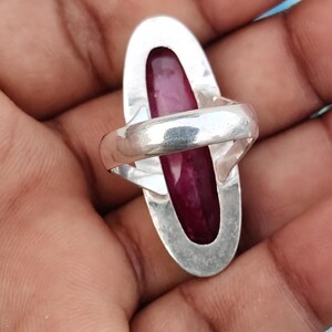 May include: A silver ring featuring a long, oval-shaped, deep magenta-colored gemstone set vertically within a smooth, polished silver frame. The band of the ring is also made of polished silver and connects to the back of the oval setting.