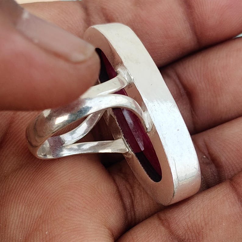 May include: A silver ring featuring a large, elongated oval-shaped deep red gemstone set within a smooth, polished metal bezel. The band consists of two separate silver wires that converge at the sides to support the prominent, vertically oriented stone.