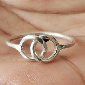 Può includere: Un anello in argento caratterizzato da due fasce circolari intrecciate. Il metallo presenta una finitura lucida e riflettente con una texture leggermente martellata sulla superficie dei due cerchi collegati, creando un design continuo.