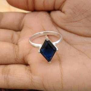 May include: A silver ring featuring a large, dark blue, diamond-shaped gemstone. The gemstone is set in a four-prong setting, and the band is a simple, polished silver. The ring is shown on a hand.