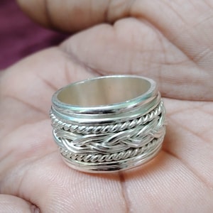 May include: A silver-colored ring with a braided design around the band. The ring has a wide band with multiple layers of braided and twisted metal, creating a textured appearance. The ring is shown on a person's hand.