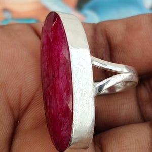 May include: A ring featuring a large, elongated, oval-shaped deep red gemstone set in a smooth, polished silver-toned metal bezel. The band of the ring is split into two separate, slender silver-toned metal arms that connect to the main setting.