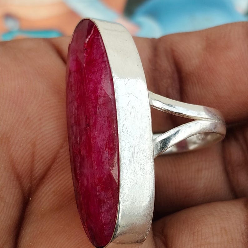 May include: A ring featuring a large, elongated, oval-shaped deep red gemstone set in a smooth, polished silver-toned metal bezel. The band of the ring is split into two separate, slender silver-toned metal arms that connect to the main setting.